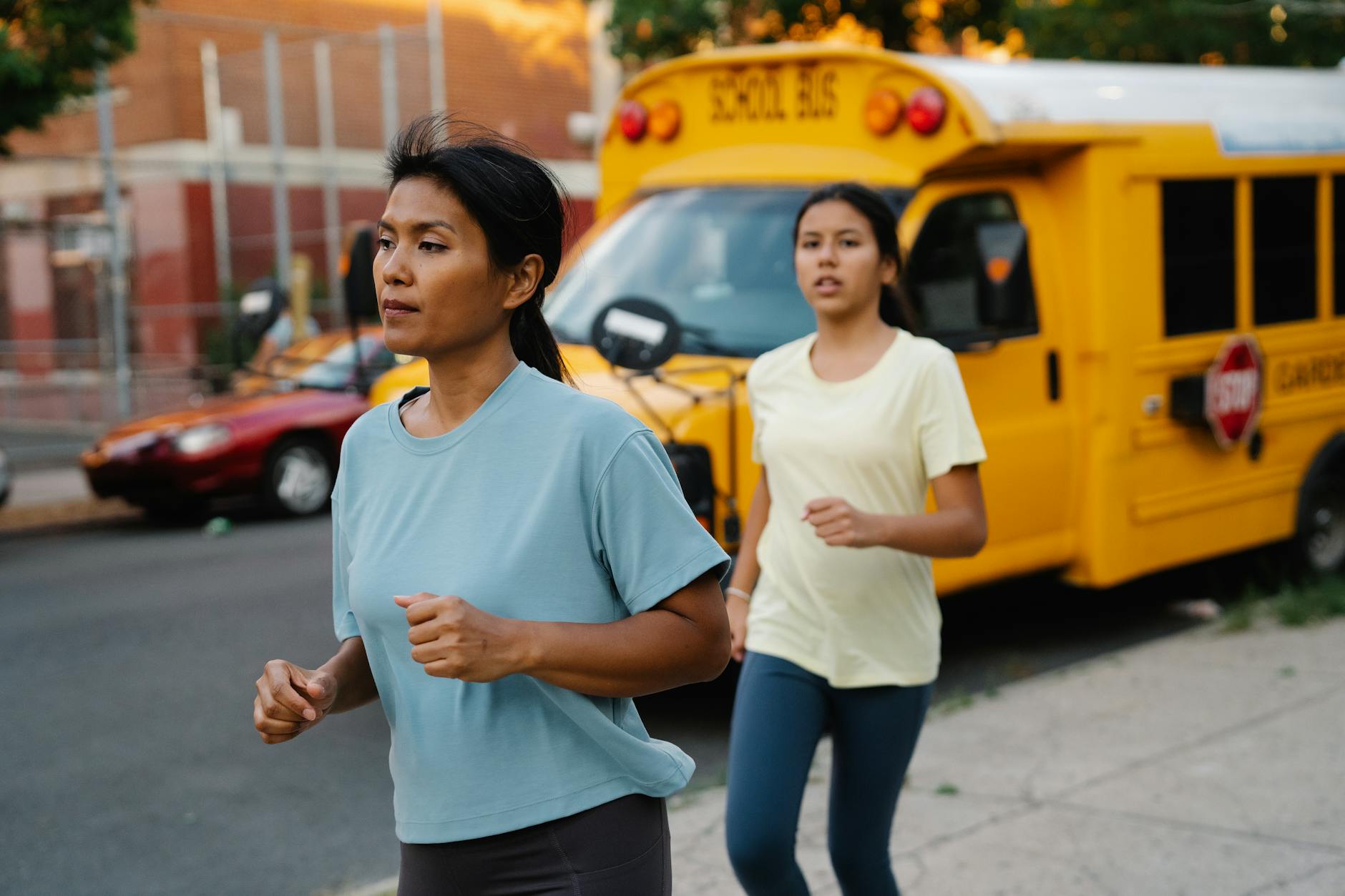A mother and daughter jogging outdoors near a parked yellow school bus on a city street.